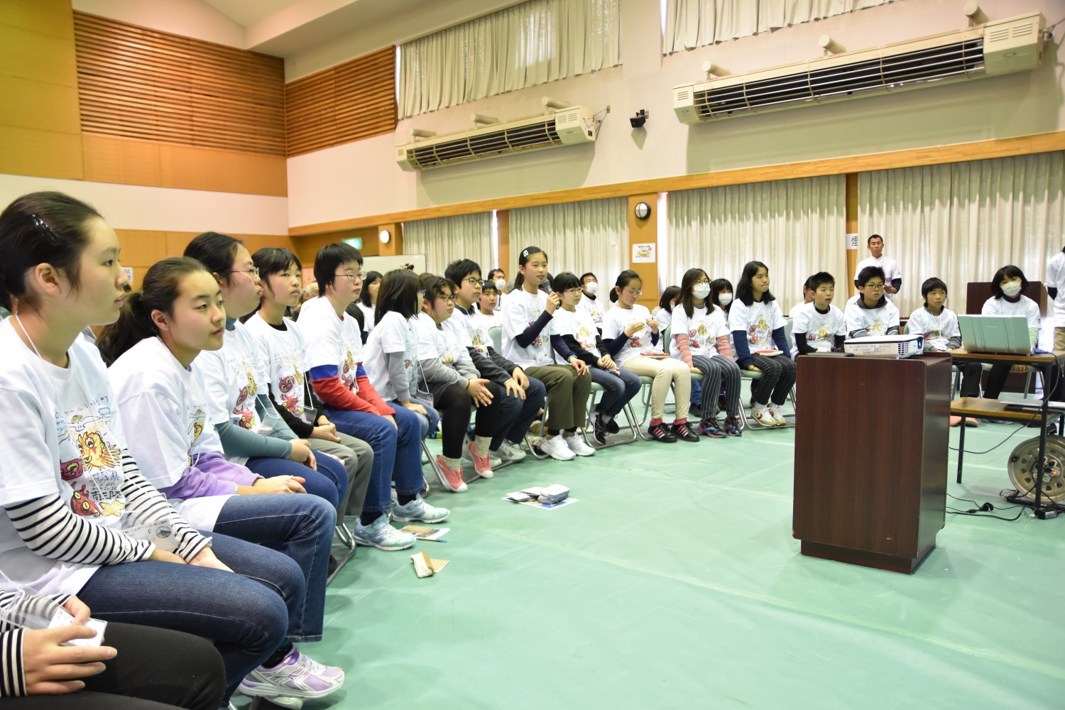 多くの子供たちが白いTシャツを着て、室内で弧を描くように並べられた椅子に座っている写真
