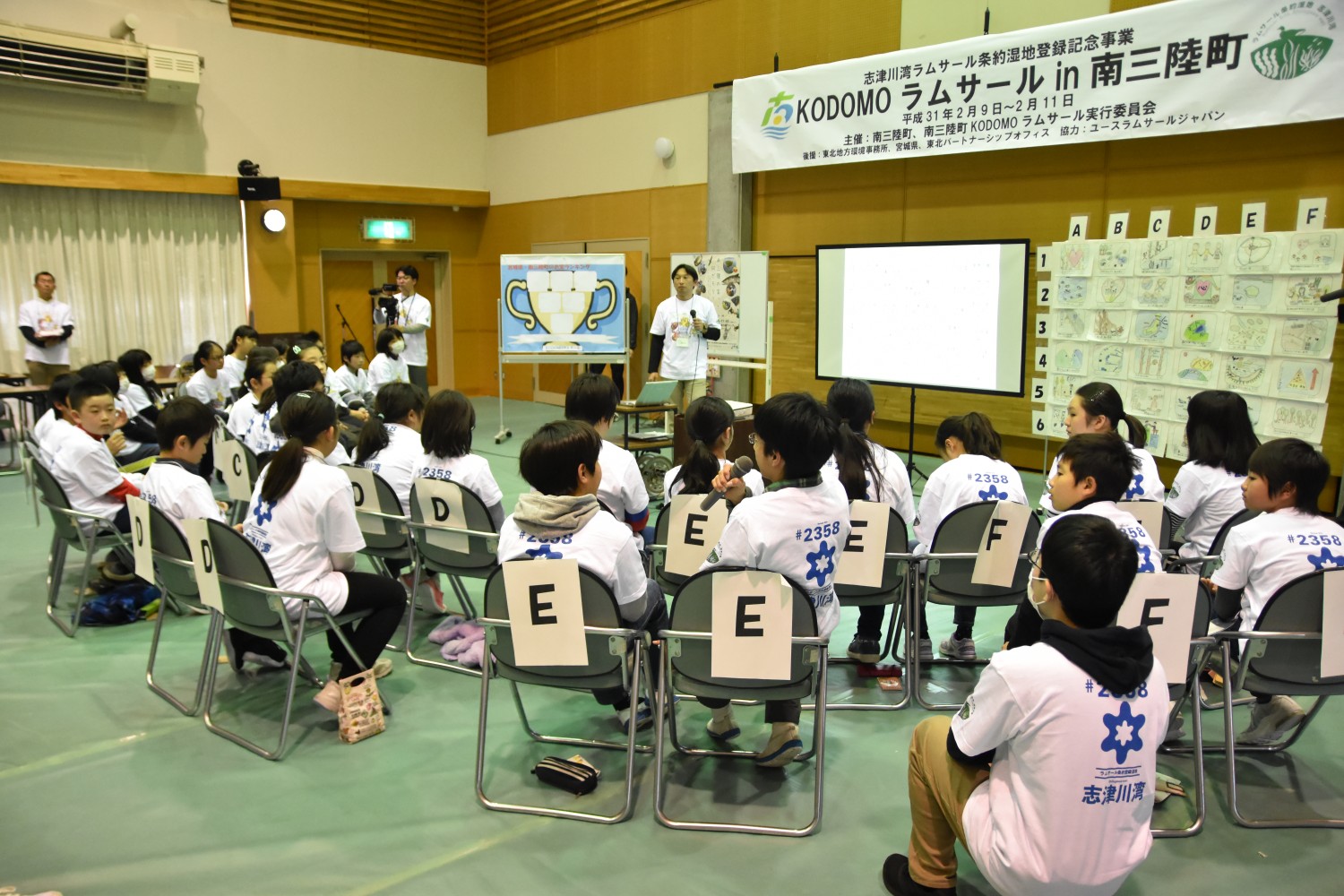 パイプ椅子が並べられた広い室内で子供たちが椅子に座って前方を見ており、壁には「KODOMOラムサールin南三陸町」と書かれた横断幕が掲げられ、ホワイトボードとプロジェクターが設置されている写真