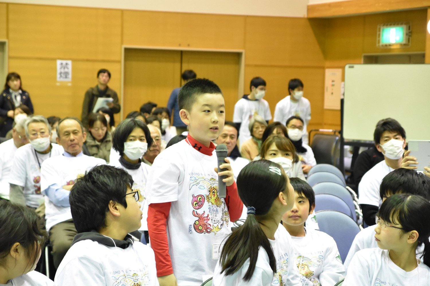 白いTシャツを着た子供たちが椅子に座っており、その中の一人が立ち上がってマイクを持ち、前に向かって話している様子が写っている写真