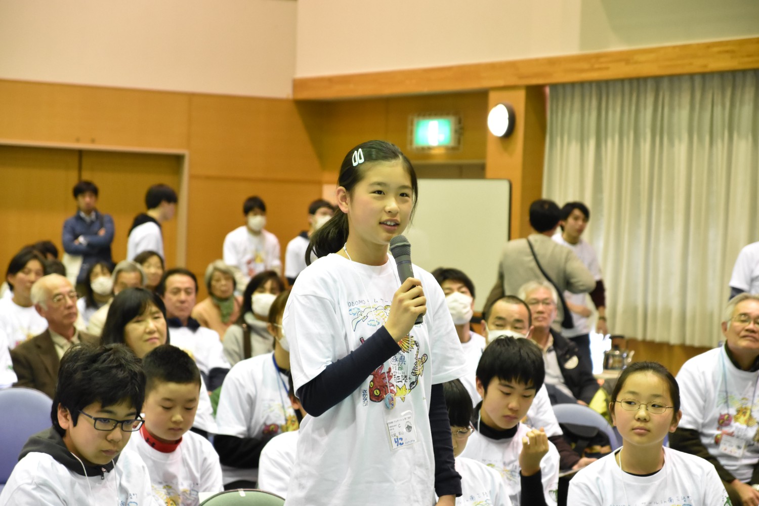 多くの人が集まっている室内で、白いTシャツを着て髪を一つにまとめた女の子が立ってマイクを持ち、発言している様子が写っている写真