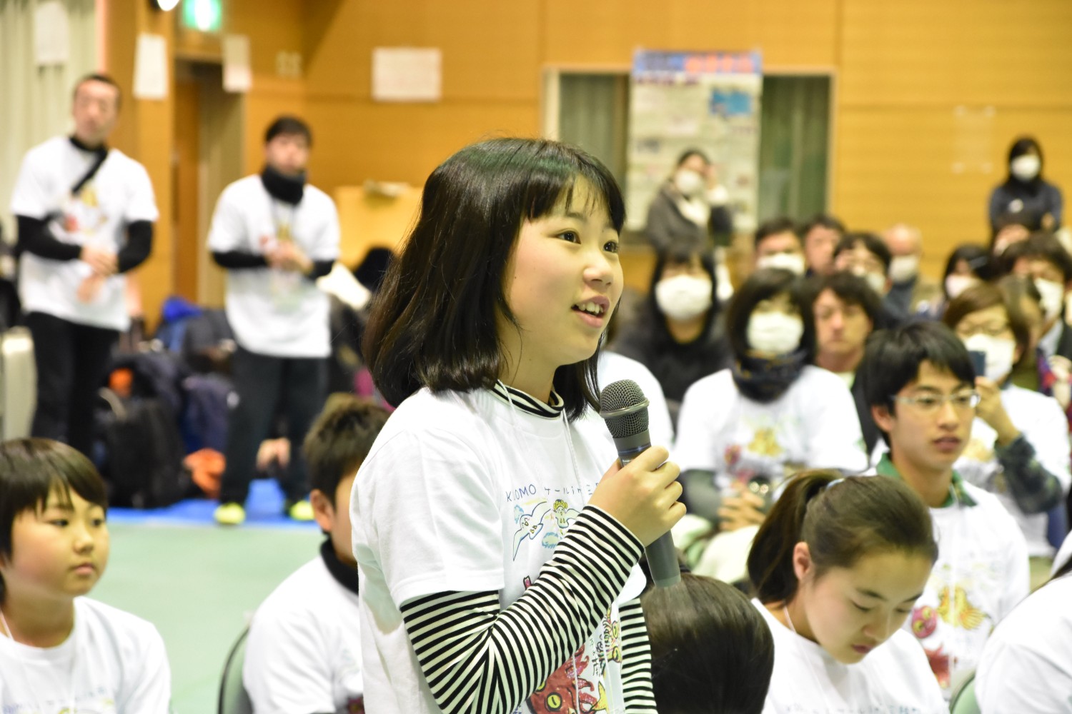 室内で多くの人が集まっている中、白いTシャツを着た女の子が立ってマイクを持ち、話している様子が写っている写真