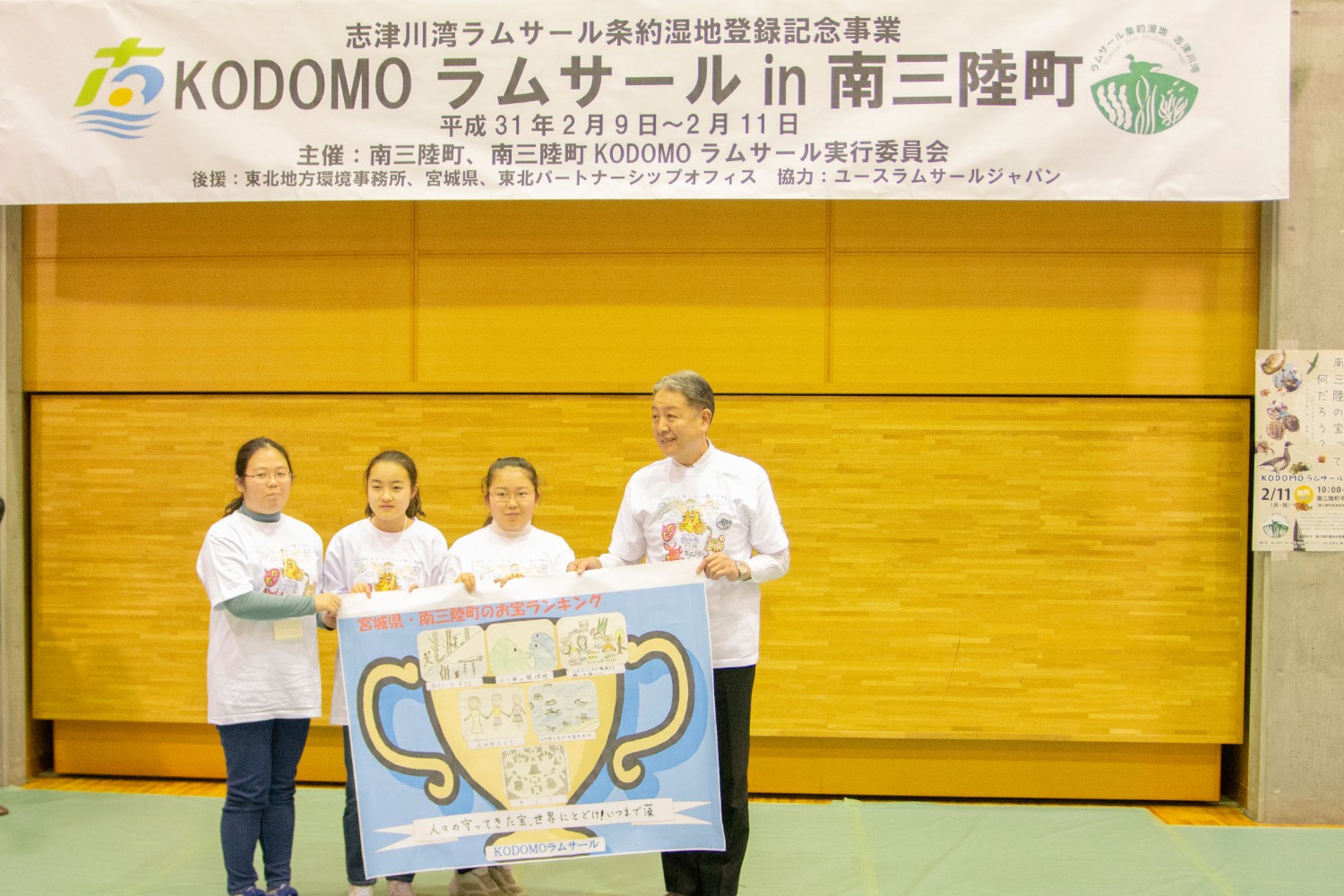 志津川湾ラムサール条約湿地登録記念事業「KODOMOラムサールin南三陸町」の横断幕が掲げられている下で、金色の大きなトロフィーの絵が描かれたボードを3人の子供たちと男性が一緒に持っている写真