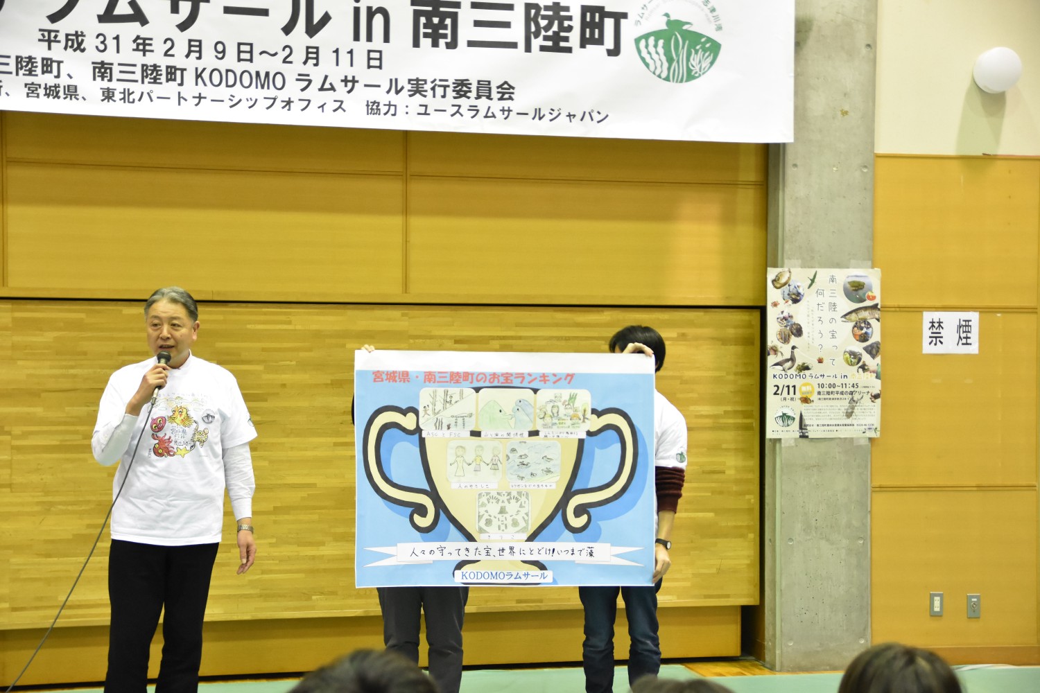 壇上で一人の男性がマイクを持って話しており、その隣で別の二人が金色の大きなトロフィーの絵が描かれたボードを両手で持っている写真