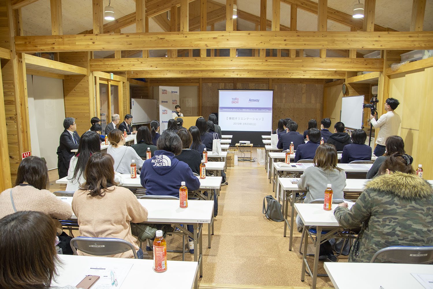 木造の会場で、多くの人が机に座って資料が映し出されたスクリーンを見ている研修の様子を後方から撮影した写真