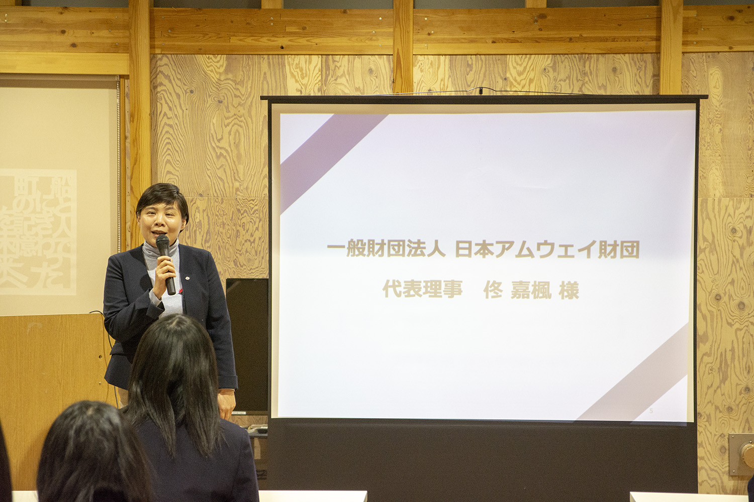 スクリーンの左側でマイクを持った女性が話をしている写真