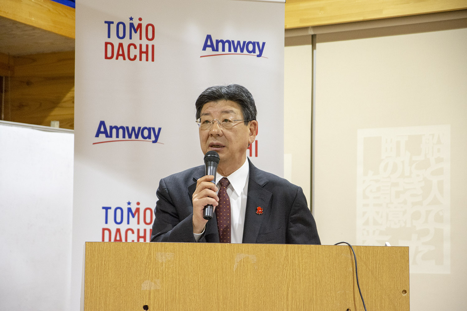 TOMODACHIとAmwayの垂れ幕の前の檀上で、マイクを持ったスーツ姿の佐藤町長が話している写真