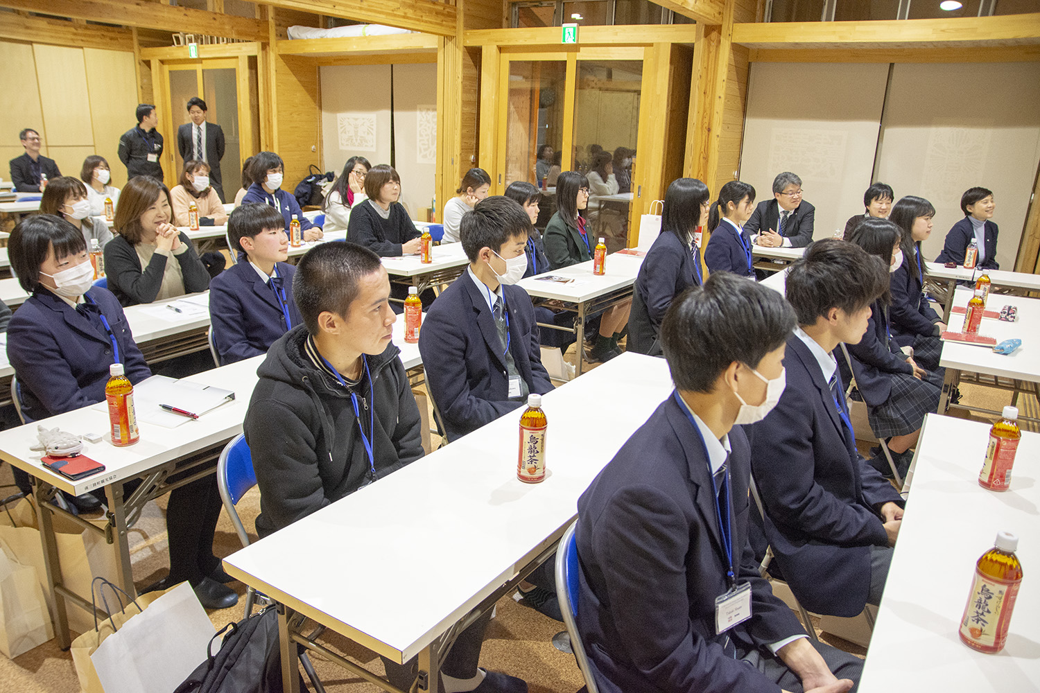 研修に参加している高校生や大人が2人ずつ机に座り、真剣に前を見つめて話を聞いている写真