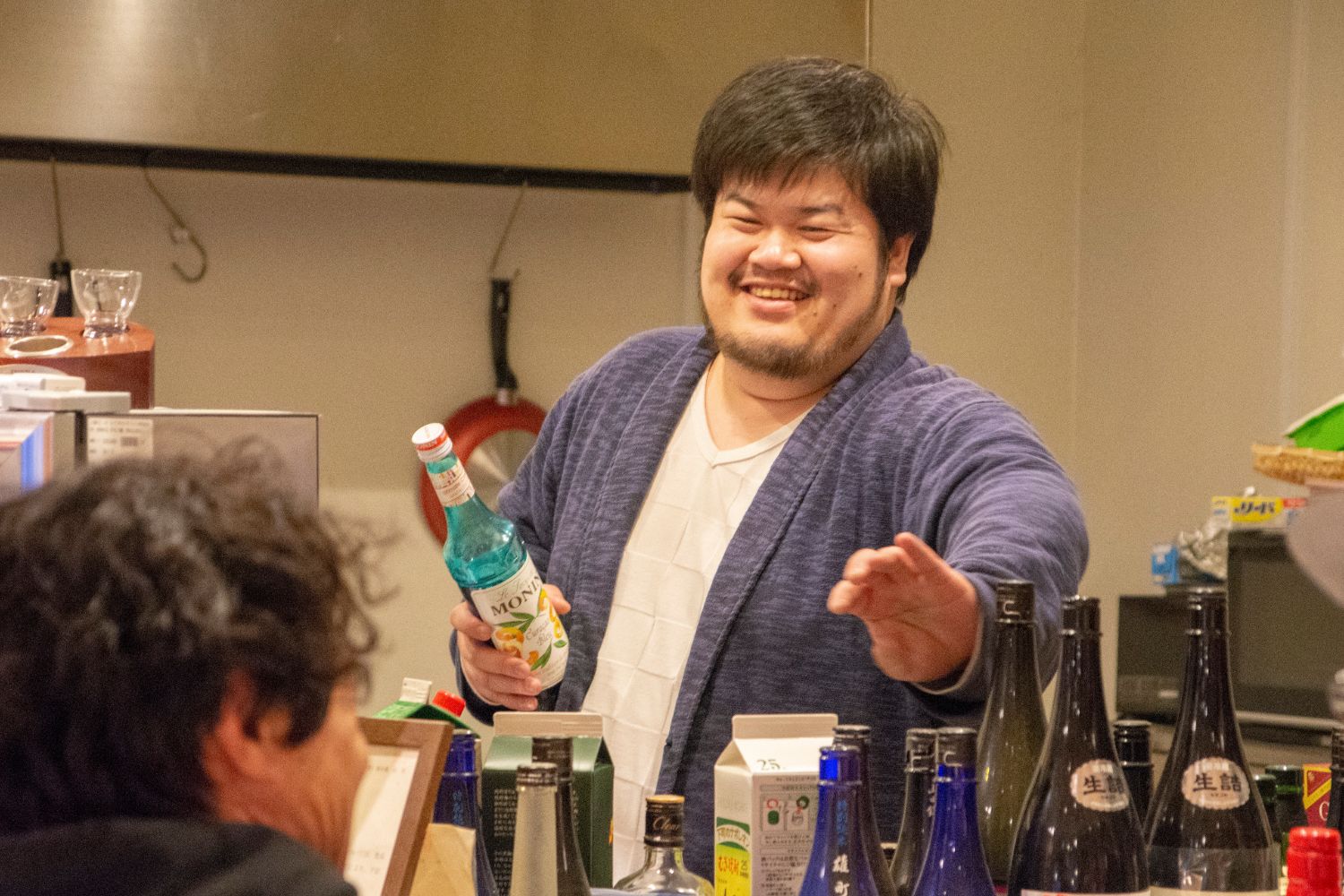 酒屋のカウンターで、笑顔の佐藤さんが青い瓶を手に取りながら笑顔で接客している写真