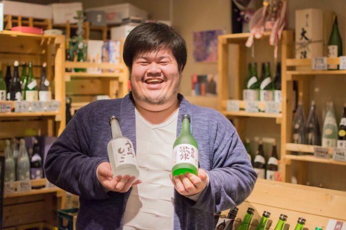 店内の木製棚にずらりと並ぶ酒瓶を背景に、薄い紫色のカーディガンを着た笑顔の佐藤裕さんが日本酒の瓶を両手に持っている写真