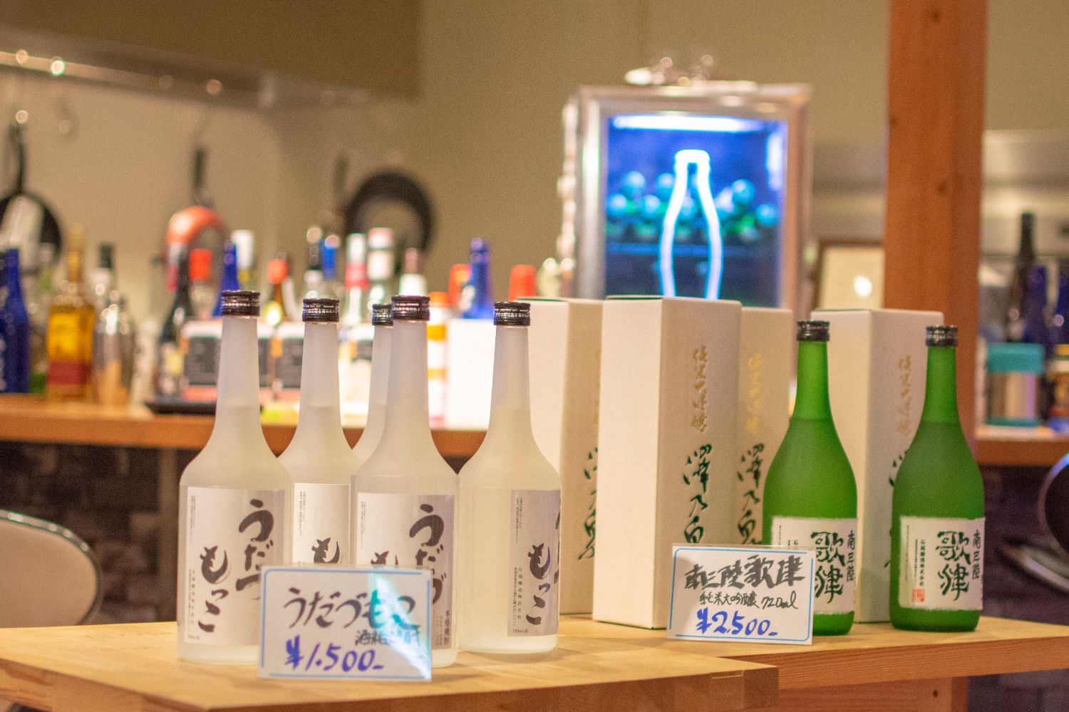 手前に値札が置かれているカウンターに、白い瓶の日本酒と緑の瓶の日本酒が整然と並んでいる写真