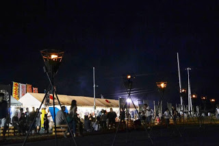 夜の屋外に設置された複数のトーチの灯りがともる中、多くの人が白いテントの周りに集まり夏祭りを楽しんでいる様子の写真