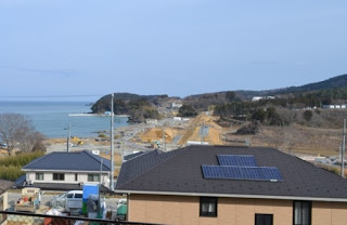 手前に屋根に太陽光パネルを載せた住宅があり、その奥に海へと続く造成中の道路と重機が見える沿岸地域の写真