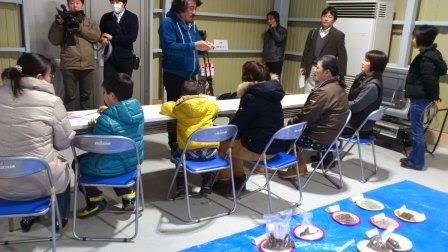 折りたたみ椅子に座った親子の参加者がテーブルに向かって並び、後方には立っている人やカメラを持った関係者がいる室内で、手前には青いシートの上に材料が乗った皿が並べられている、ガラス玉作成のワークショップの様子の写真