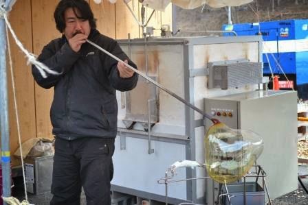 ガラス工房で、長い金属の吹き竿の先にある溶けたガラスを成形している村山耕二さんが、炉の前で作業しているガラス吹き作業中の写真