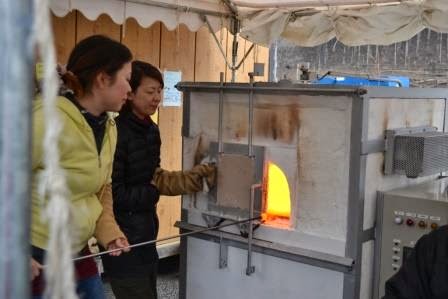 黄色い上着を着た女性参加者が長い棒を持ち、女性スタッフが開けた炉の扉からは強いオレンジ色の光が見える、ガラス玉作り体験中の様子を捉えた写真