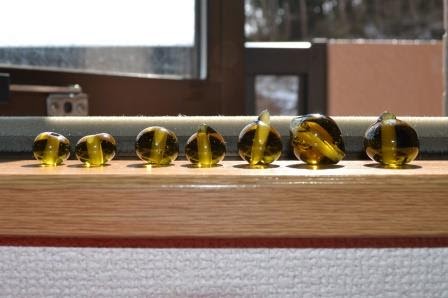 木製の台の上に、縦中央が黄色で黒色の大小様々な7つのビー玉が一列に並べられ、窓から差し込む日差しがビー玉を照らしている写真