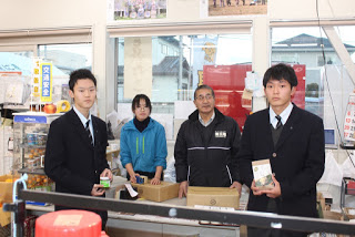 男性と女性が並んで立っているお店のカウンターに、2つの段ボールの小箱が置かれており、その手前に商品を手に持った制服姿の男子学生が2人カメラを向いて立っている写真