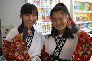 赤い花柄の袖が特徴的な華やかな伝統衣装を着た中学生の女子二人が、笑顔でピースサインをして写っている写真