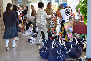 会場のロビーで、中学生と大人たちが文化祭の準備をしており、鹿子躍の鹿の角飾りが入った大きな袋のそばで伝統衣装の着替えや荷物の整理をしている様子を写した写真