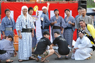 浴衣を着た男性たちが赤白の幕の前で笑顔を見せ「南三陸ふっこう青年会」のメンバーが写っている写真