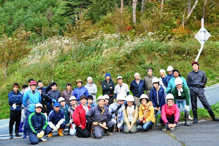 参加者たちが道路脇に集まり、作業服姿で笑顔を見せながら記念撮影をしている様子の写真