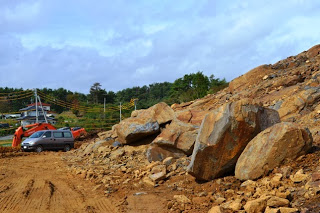 掘削された斜面に大小の岩が転がり、近くに重機と車が停まっている写真