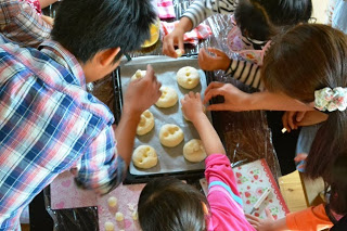 女の子2人と大人の男女がオーブン用天板に並べた丸いパン生地に手を伸ばして飾りつけしている様子を上から写した写真
