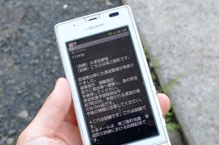 携帯電話の画面に緊急速報メールの文面を開いて見せている写真