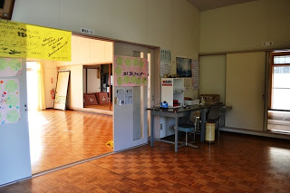 木目調の床が広がる明るい室内で、左側の開いた引き戸の奥には集会所のような広いスペースがあり、入口の壁には手書きのポスターや案内が貼られている地域施設内の写真