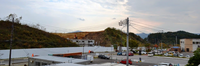 小高い丘が削られ、白い囲いのある工事現場の向こうに駐車場と建物が並んでいる写真