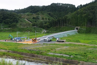 緑の田園が広がる中、トンネルへと続く道路が建設され、工事車両が作業を行っている写真