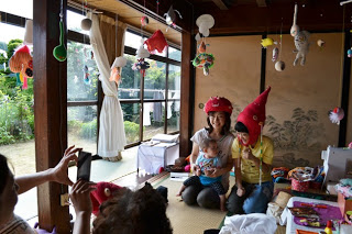 畳の部屋の中で赤い帽子をかぶった女性二人が赤ちゃんをあやしながら笑顔で写真を撮られており、天井からは色とりどりのアート作品が吊るされている様子の写真