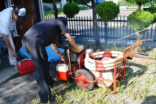 黒い服を着た男性が赤い小型の可搬式ポンプに手をかけてエンジン部分を確認しており、そばには燃料缶やホースが載っていて、もう一人の男性が見守る中で災害時の備品の動作点検を行っている写真