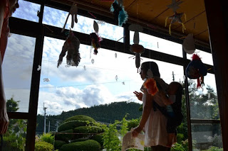 赤ちゃんをおんぶした女性が大きく開いたガラス戸から、外の手入れされた庭と先に広がる緑の景色を見ており、室内にはたくさんのアート作品が吊るされている様子の写真