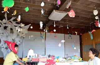 和室の天井からカラフルな吊り飾りがたくさん下がっており、赤い帽子をかぶった女性と年配の女性が向かい合って話しているアート展示会場の写真