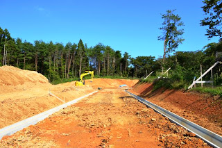 広くならされた土地の両側にコンクリートの溝が作られ、水が流れるように整えられており、重機も見える道路づくりの途中の写真