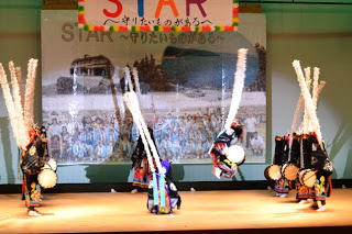 太鼓を腰に付けた踊り手たちが、本物の鹿角を模した頭飾りと白い長い竹飾り（ササラ）を背負い、黒を基調とした衣装に五色の布をなびかせながら舞台の上で円を描くように踊っている、行山流水戸辺鹿子躍の写真