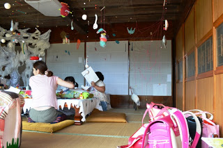 畳敷きの和室で小学生たちがテーブルを囲んで糸や布などの素材に触れながらアートづくりを楽しんでおり、天井からは手作りの吊り飾りがぶら下がり、手前にはランドセルが置かれていて、放課後にアートイベントに訪れた子どもたちの様子が伝わる写真