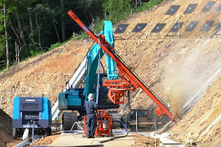 作業員が重機と掘る機械を使って斜面に斜めの穴をあけ、斜面を安定させる工事をしている様子を写した斜面補強工事中の写真