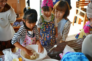エプロンをつけた女の子が白いボウルで生地を混ぜているのを、エプロンをつけた小さな女の子と女性が一緒に見守っている料理教室の様子の写真