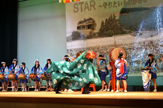 獅子舞が舞台中央で力強く舞い、後方では青い法被を着た和太鼓担当の生徒たちが並んでいる様子を写した写真