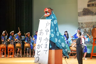 ステージ中央で獅子舞の装束を着た人物が「戸中 今までありがとう」と書かれた横断幕を掲げており、後方では法被姿の生徒たちが太鼓を演奏している様子を写した写真