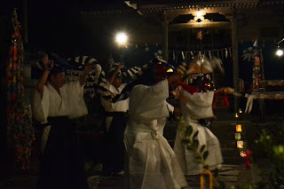 神社の境内で白い上着に袴を着けた神楽の舞手たちが、鈴や扇を手に持ち、夜の灯りの中で舞を奉納している様子の写真