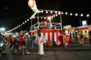 夜の夏祭り会場で紅白の幕がまかれたやぐらの周囲を浴衣姿の人々が囲み、提灯が灯る中で盆踊りが行われている様子の写真