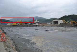 背景には曇り空と山々が広がり、泥でぬかるんだ地面の上に複数のショベルカーやトラックが並ぶ現場の左中央が赤枠で囲まれている工事中の写真