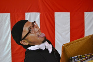 赤白の幕の前で、眼鏡と帽子をかぶり、白いタオルを首に巻いた男性が大きな段ボールを持ちながら舌を出してユーモラスな表情を見せている男性の写真