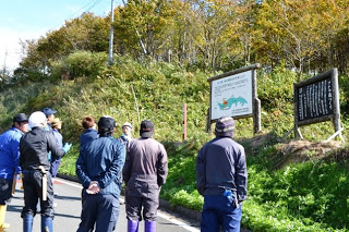 作業服を着た参加者たちが道端に設置された「ブナの森づくり」の案内看板の前に集まり、説明を聞いている様子の写真