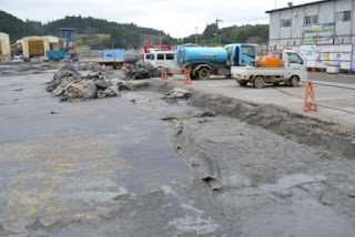 青い給水車やオレンジ色のタンクを積んだ白いトラックなどの車両が停まり、左側のぬかるんだ地面にシートが敷かれ、オレンジ色のバリケードが配置された作業中の工事現場の写真