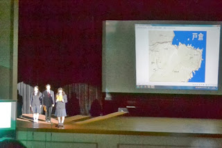 制服姿の生徒3人が舞台に立ち、大型スクリーンに「戸倉」と書かれた地図を映しながら、地域についての発表をしている様子を写した写真