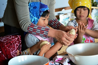 子ども二人がカラフルなバンダナを頭に巻き、大人に手伝われながら生地を丸めて成形する体験をしている様子の写真